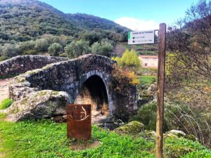Puente de piedra medieval en el Camino de los Trasiegos con señalización turística y montañas de la Sierra de Francia.