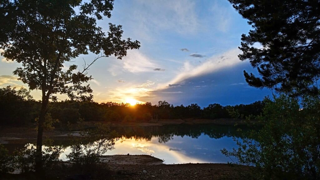 Atardecer en lago tranquilo rodeado de árboles verdes en la Sierra de Francia, cielo azul con nubes doradas