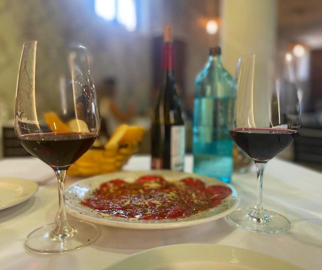Mesa de comedor con dos copas de vino tinto, plato de jamón ibérico y botellas de vino al fondo en una cena.