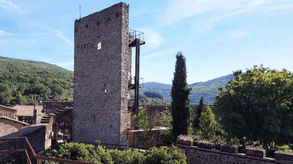 Torre medieval de piedra con cruz metálica en Sierra de Francia, rodeada de vegetación y montañas boscosas al fondo.