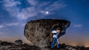 Hombre sostiene enorme roca bajo cielo estrellado en la Sierra de Francia durante la noche.