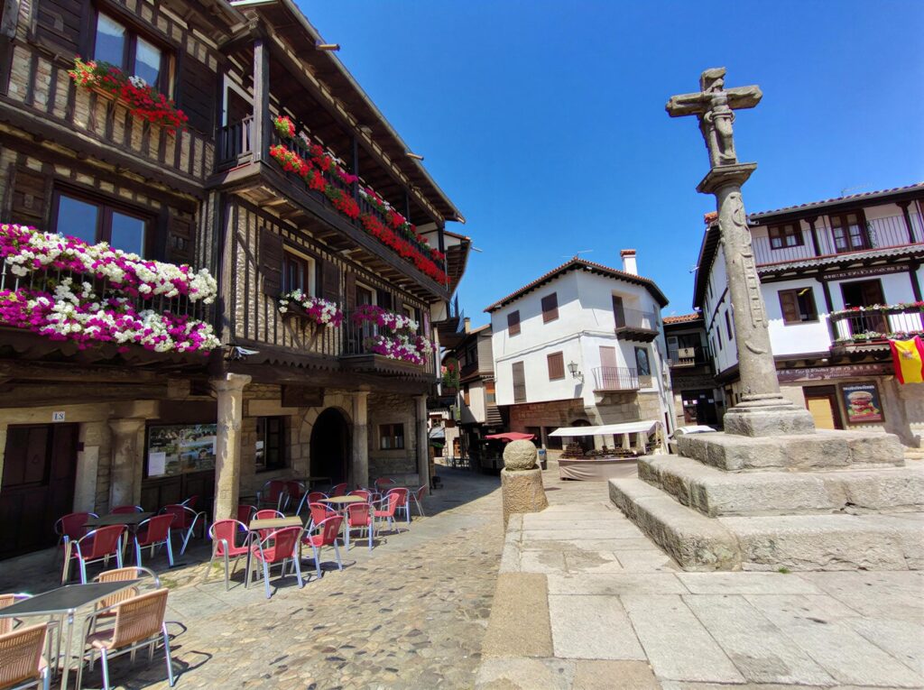 Plaza mayor de La Alberca con casas tradicionales, cruz de piedra y flores decorativas en día soleado.
