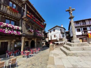 Plaza mayor de La Alberca con casas tradicionales, cruz de piedra y flores decorativas en día soleado.