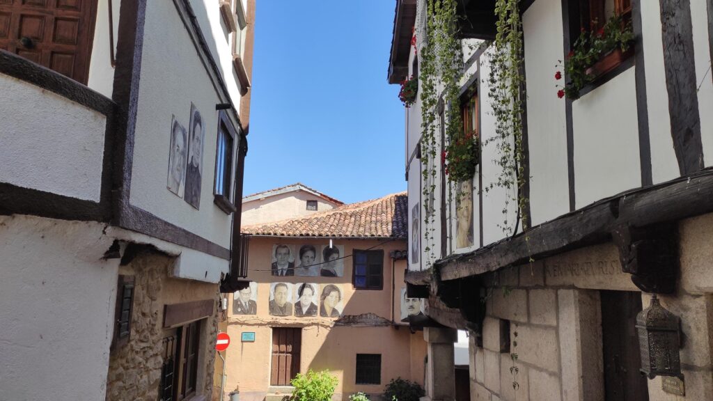 Calle empedrada de Mogarraz con casas blancas, retratos en fachada y vegetación colgante bajo cielo azul.