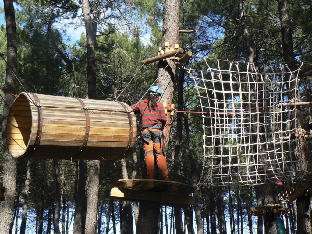 Persona en arnés atravesando tubo de madera en circuito de aventura entre árboles del bosque.
