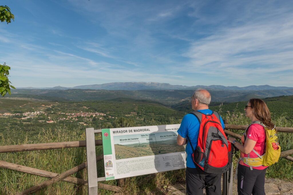 Dos senderistas leen panel informativo en Mirador de Madroñal, Sierra de Francia, con vistas a valle y montañas nevadas