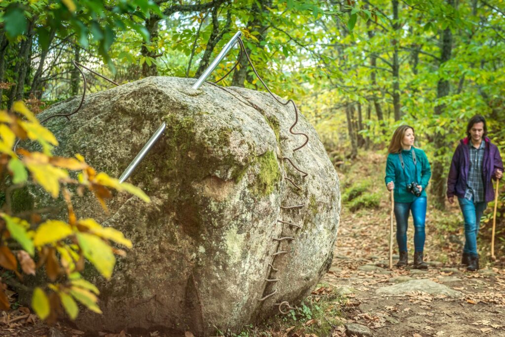 Dos jóvenes senderistas caminan junto a una escultura de piedra con espejos en la Sierra de Francia.