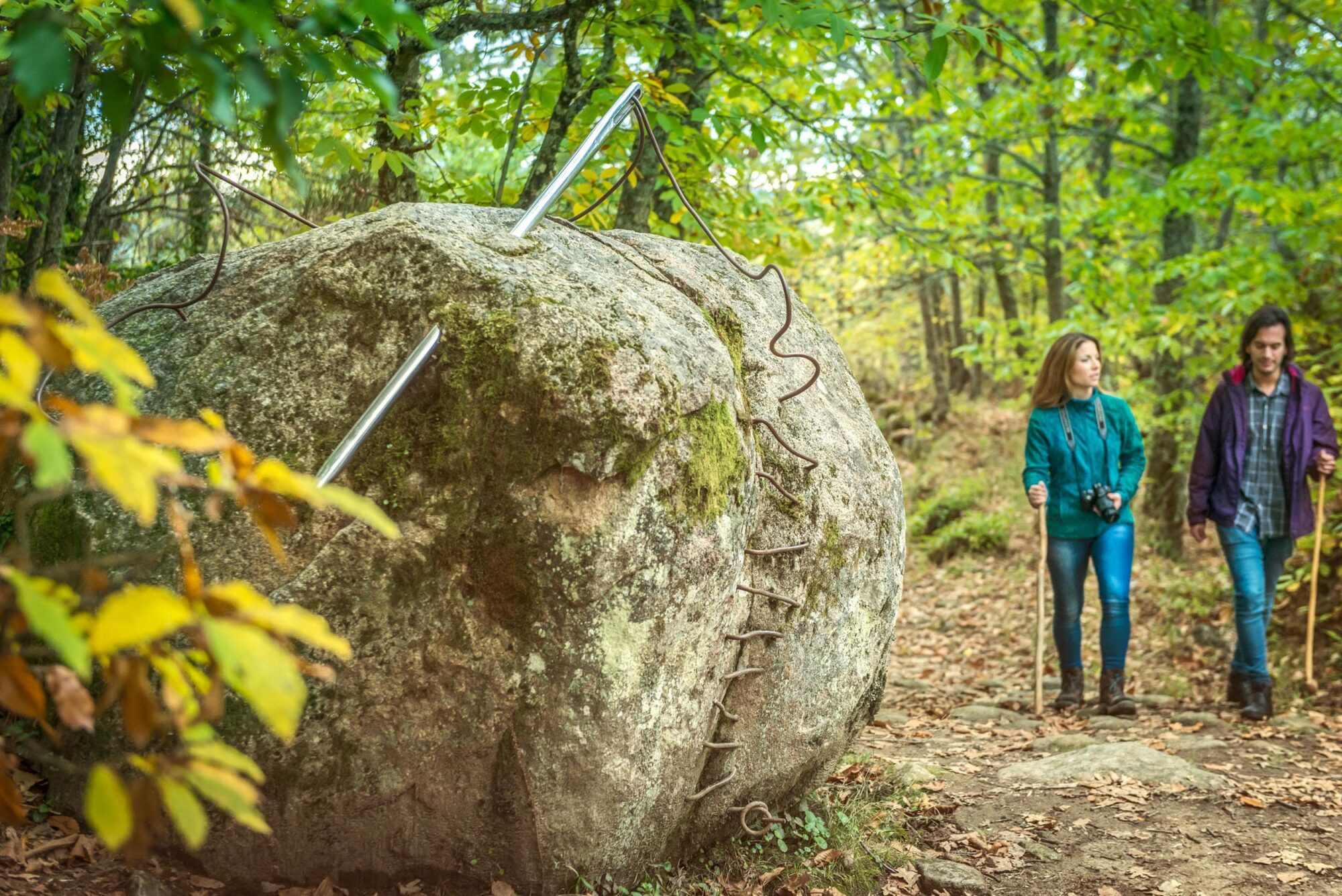 Dos jóvenes senderistas caminan junto a una escultura de piedra con espejos en la Sierra de Francia.