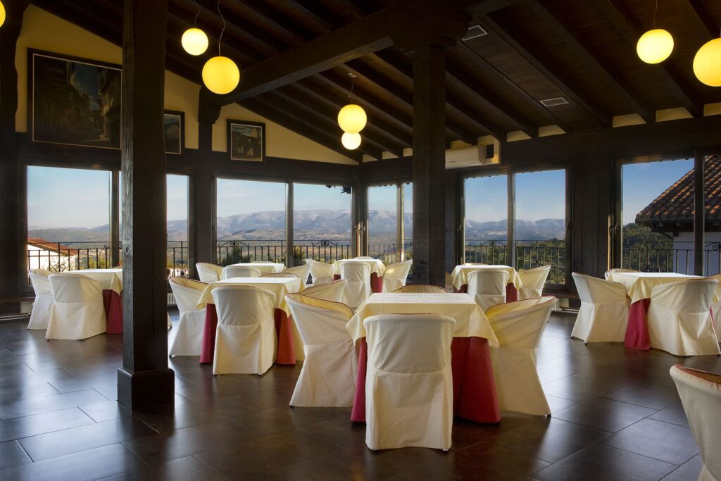 Salón elegante con vistas panorámicas a montañas, mesas redondas decoradas en blanco y rojo, iluminación cálida.