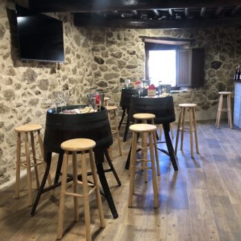 Bodega El Abuelo Flores: interior rústico con paredes de piedra, mesas altas negras y taburetes de madera clara para degustación de vinos.