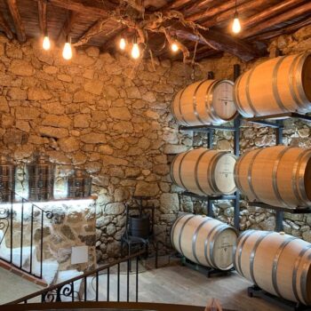 Bodega tradicional con barriles de madera en estanterías metálicas, paredes de piedra y lámparas colgantes de diseño rústico.
