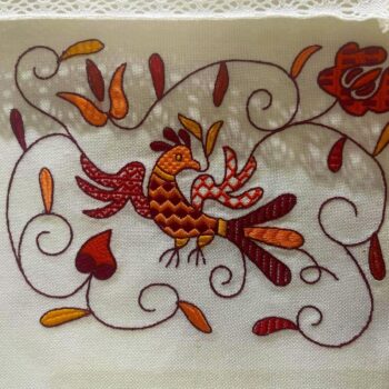 Bordado tradicional con gallo decorativo y motivos florales en tonos rojos y naranjas sobre lino blanco.