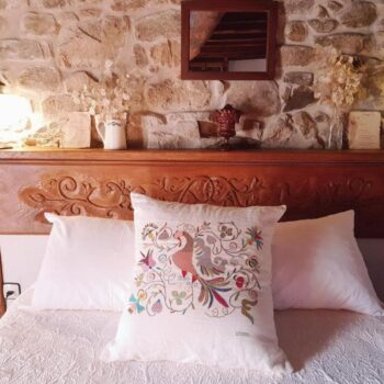 Dormitorio acogedor con cama de madera tallada, almohadas blancas, pared de piedra rústica y decoración tradicional de la Sierra de Francia.