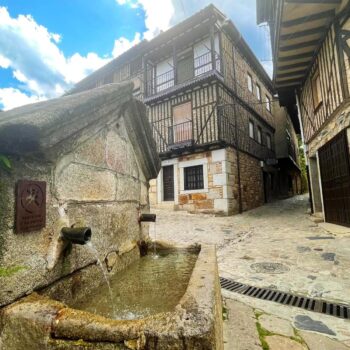 Calle empedrada de La Alberca con casas tradicionales de piedra, balcones de madera y fuente medieval bajo cielo azul.
