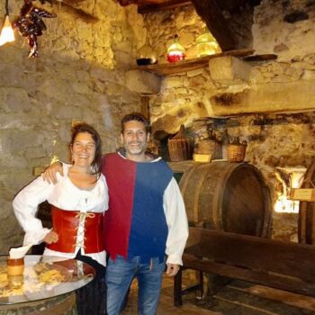 Pareja en traje medieval dentro de bodega histórica con barriles de madera y paredes de piedra antigua.