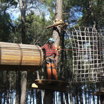 Persona en arnés atravesando tubo de madera en circuito de aventura entre árboles del bosque.