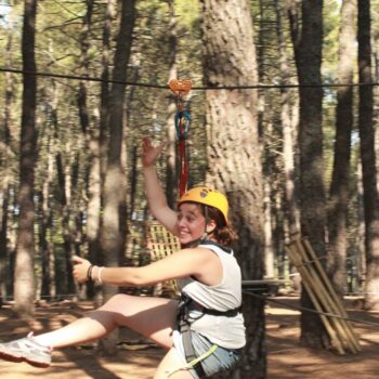 Niña en tirolesa en bosque de pinos, equipada con casco amarillo y arnés de seguridad en parque de aventura.