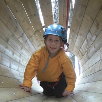 Niño con casco azul gateando dentro de tubo de madera en parque de aventura, sonriendo a cámara.