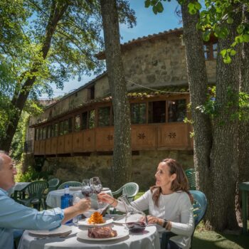 Pareja brindando en terraza sombreada de restaurante rural con vistas a arquitectura medieval de piedra entre árboles.