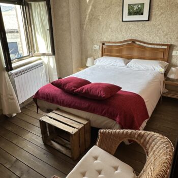 Habitación acogedora de hotel rural con cama de madera, sábanas blancas, cojines rojos y sillón de mimbre junto a ventana.