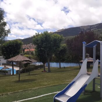 Parque infantil con tobogán azul y blanco frente a lago, montañas boscosas y cabaña de paja al fondo.
